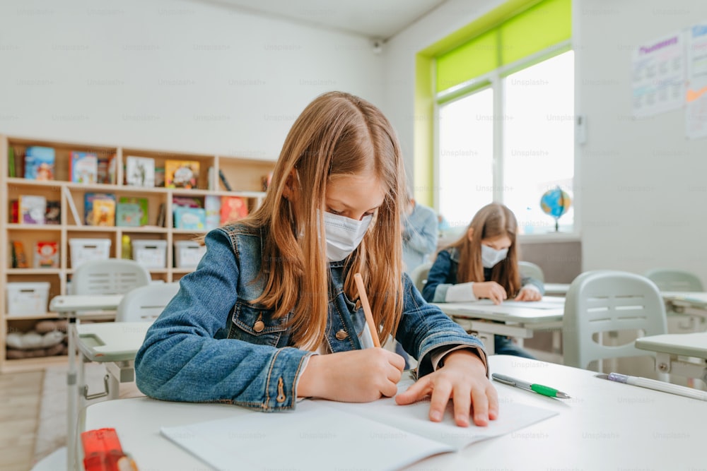 Girl in face masks solving the exercises in the school. Elementary school students during COVID-19 pandemic.