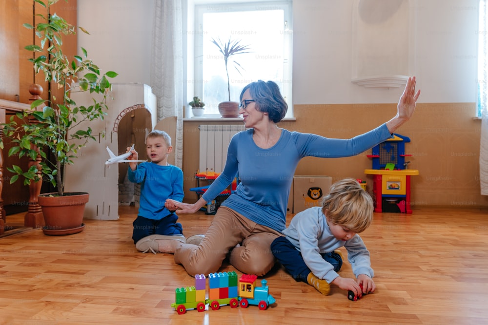 Grandmother having fun time with her grandchildren in the kids room indoor. Mature woman playing with little boys 3 and 6 years old sitting on the floor. Selective focus.
