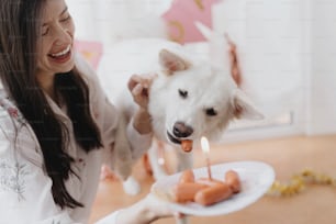 Fête d’anniversaire de chien. Jeune femme heureuse et chien affamé célébrant l’anniversaire avec un gâteau à la saucisse et une bougie sur fond de guirlande rose. Adorable berger suisse blanc premier anniversaire