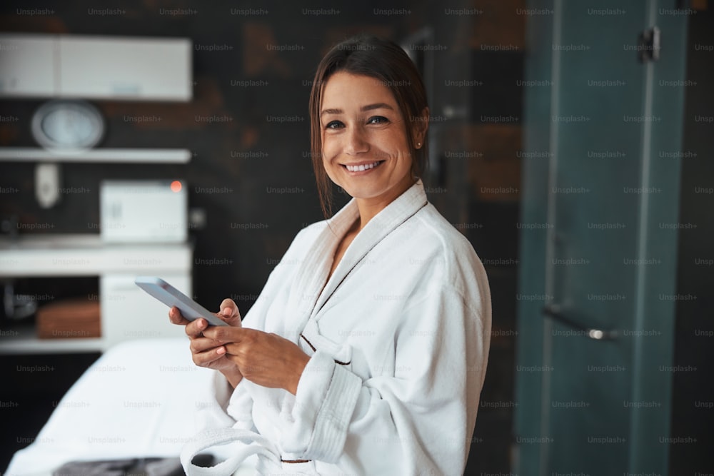 Close up portrait of happy smiling elegant female in white silk bathrobe typing message on mobile phone while standing in spa massage room
