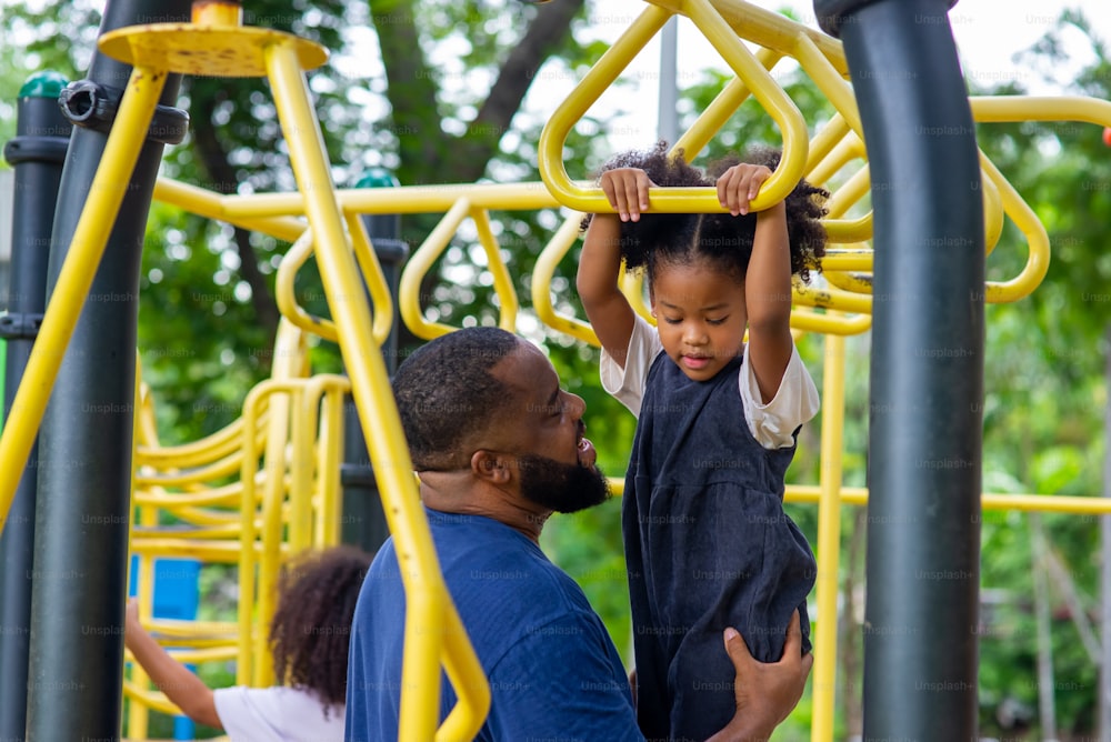 공원에서 행복한 혼혈 가족. 아프리카 아버지는 공공 공원의 어린이 놀이터에서 놀고 있는 어린 딸을 돌보고 있습니다. 아빠와 귀여운 아이 소녀 아이가 함께 야외 주말 활동을 즐기고 즐거운 시간을 보내고 있습니다.