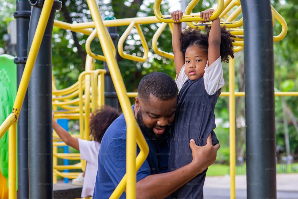 공원에서 행복한 혼혈 가족. 아프리카 아버지는 공공 공원의 어린이 놀이터에서 놀고 있는 어린 딸을 돌보고 있습니다. 아빠와 귀여운 아이 소녀 아이가 함께 야외 주말 활동을 즐기고 즐거운 시간을 보내고 있습니다.