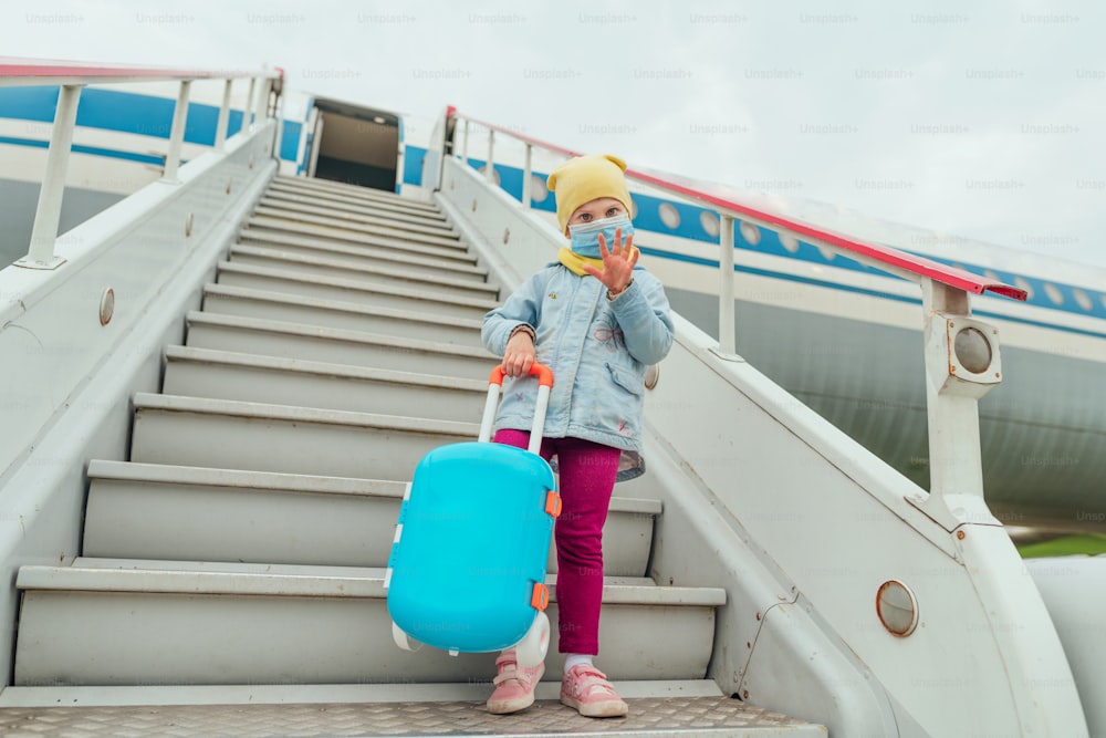 Little girl wearing protective face mask holding suitcase waving hand near the plane. Travel and tourism after the end of lockdown. New normal after Pandemic COVID-19 concept. Selective focus.