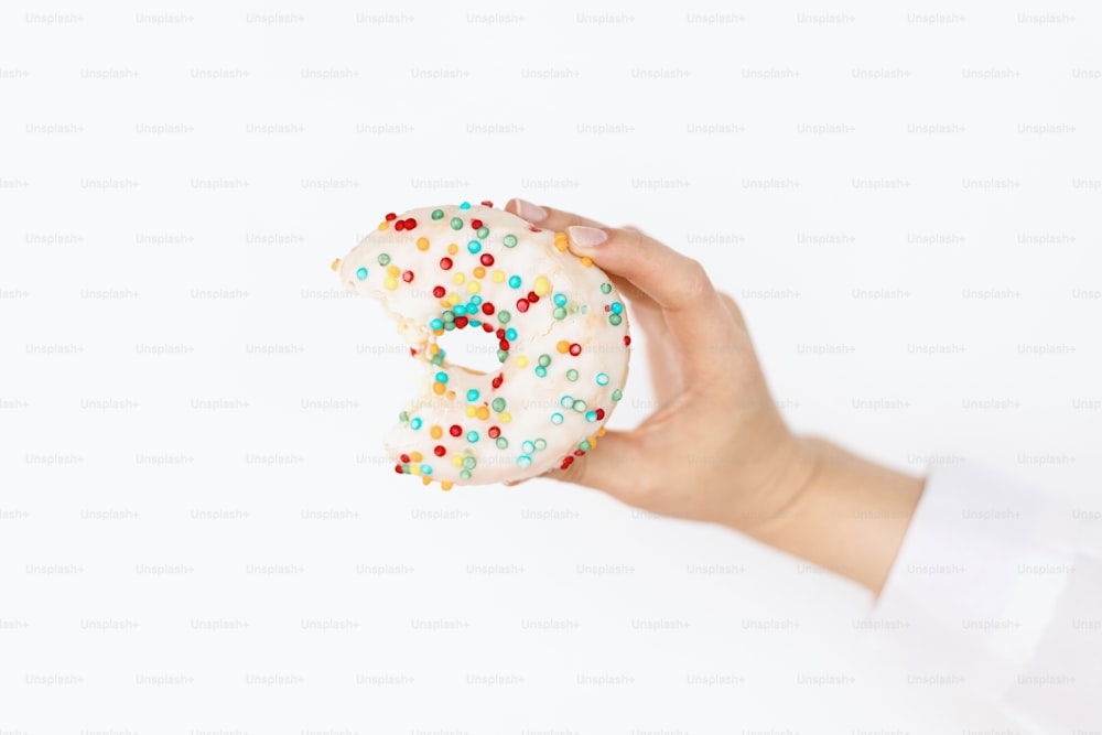 Hand holding bitten delicious donut on background of white wall in room. Birthday party celebration. Colorful doughnut with sprinkles. Diet and unhealthy food