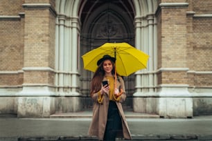 스마트폰을 사용하고 비오는 날 cit에서 걷는 동안 노란 우산을 들고 있는 젊은 아름다운 여자