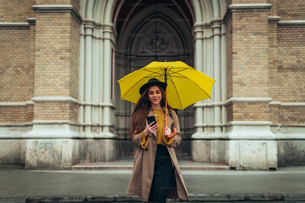Young beautiful woman using a smartphone and holding a yellow umbrella while walking in a cit on a rainy day