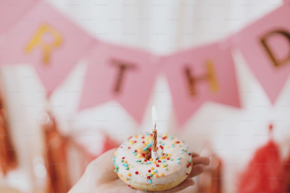 Happy Birthday. Hand holding delicious birthday donut with candle on background of pink garland and decorations in room.  Birthday party celebration. Colorful doughnut with sprinkles