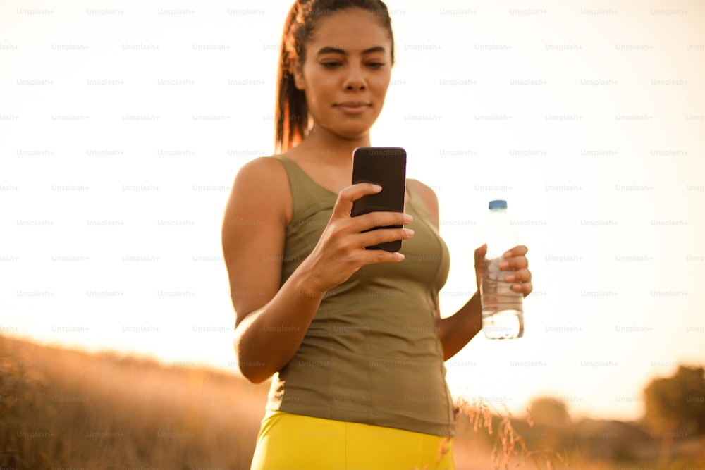 Sportliche junge Frau, die eine Flasche Wasser hält und ein Handy benutzt. Der Fokus liegt auf der Hand.