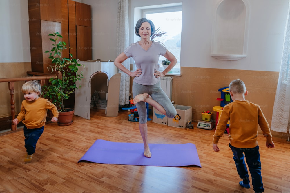Abuela activa tratando de hacer yoga mientras sus nietos corren a su alrededor en la habitación de los niños. Enfoque selectivo.