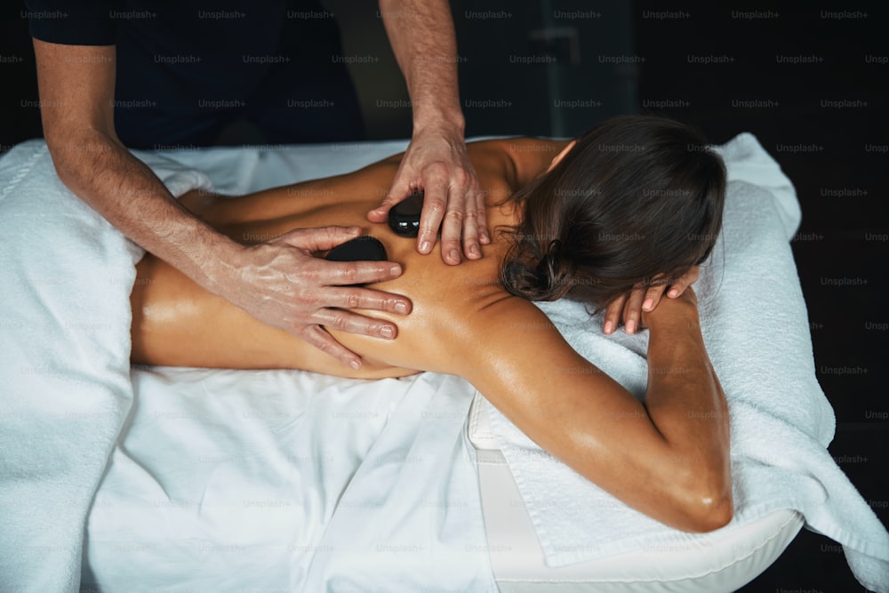Young woman lying on white towel feeling great while male therapist stroking client body with black mineral stones in spa salon
