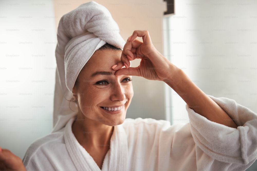 Wait up side view portrait of happy smiling Caucasian female in white bathrobe touching her skin on face in room indoors