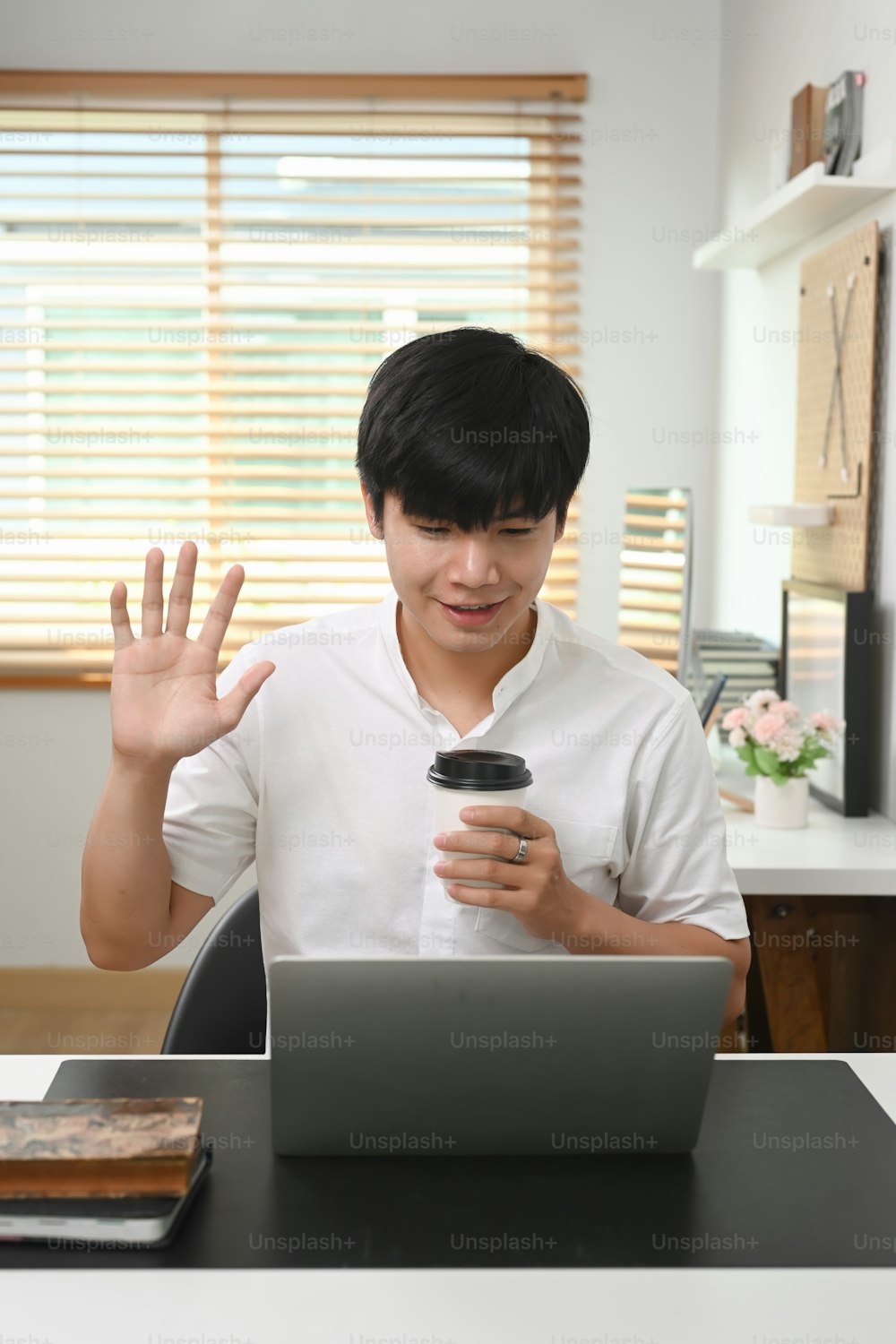 Happy young asian man holding coffee cup and greeting his friends on video call on laptop computer.