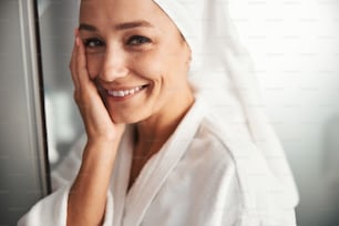 Close up side view portrait of happy adorable Caucasian woman in white bathrobe holding arm on her face while posing at the photo camera