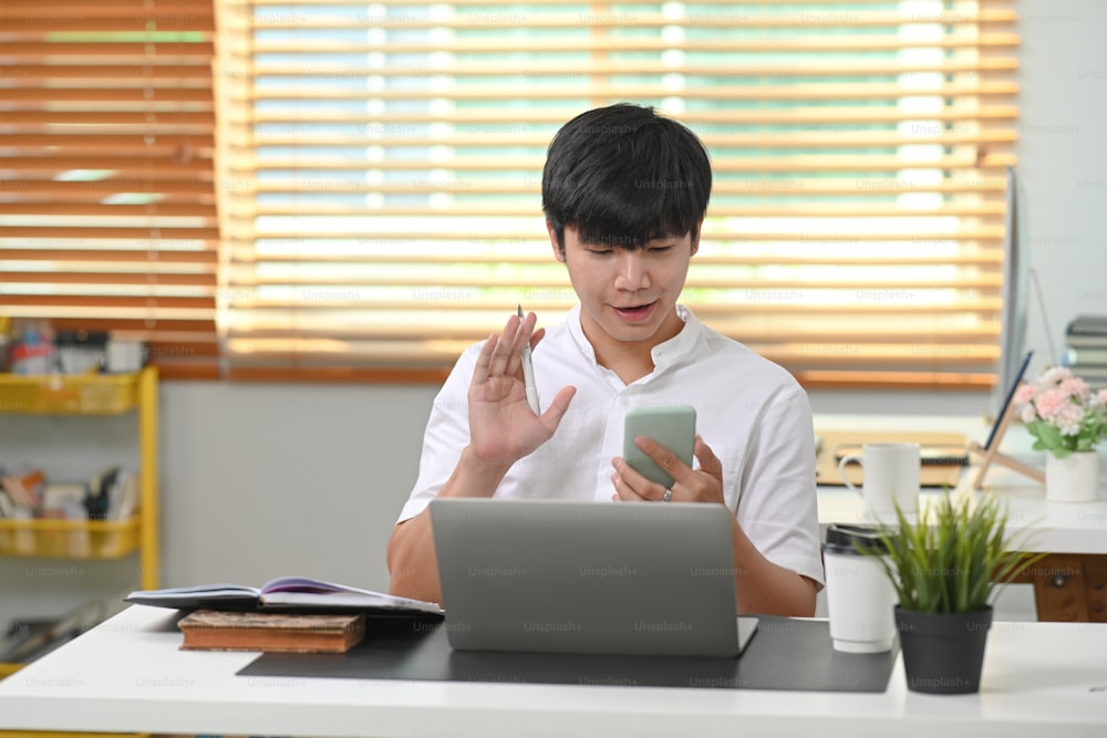 Young asian man sitting in modern home office an having video call on smart phone his friends.