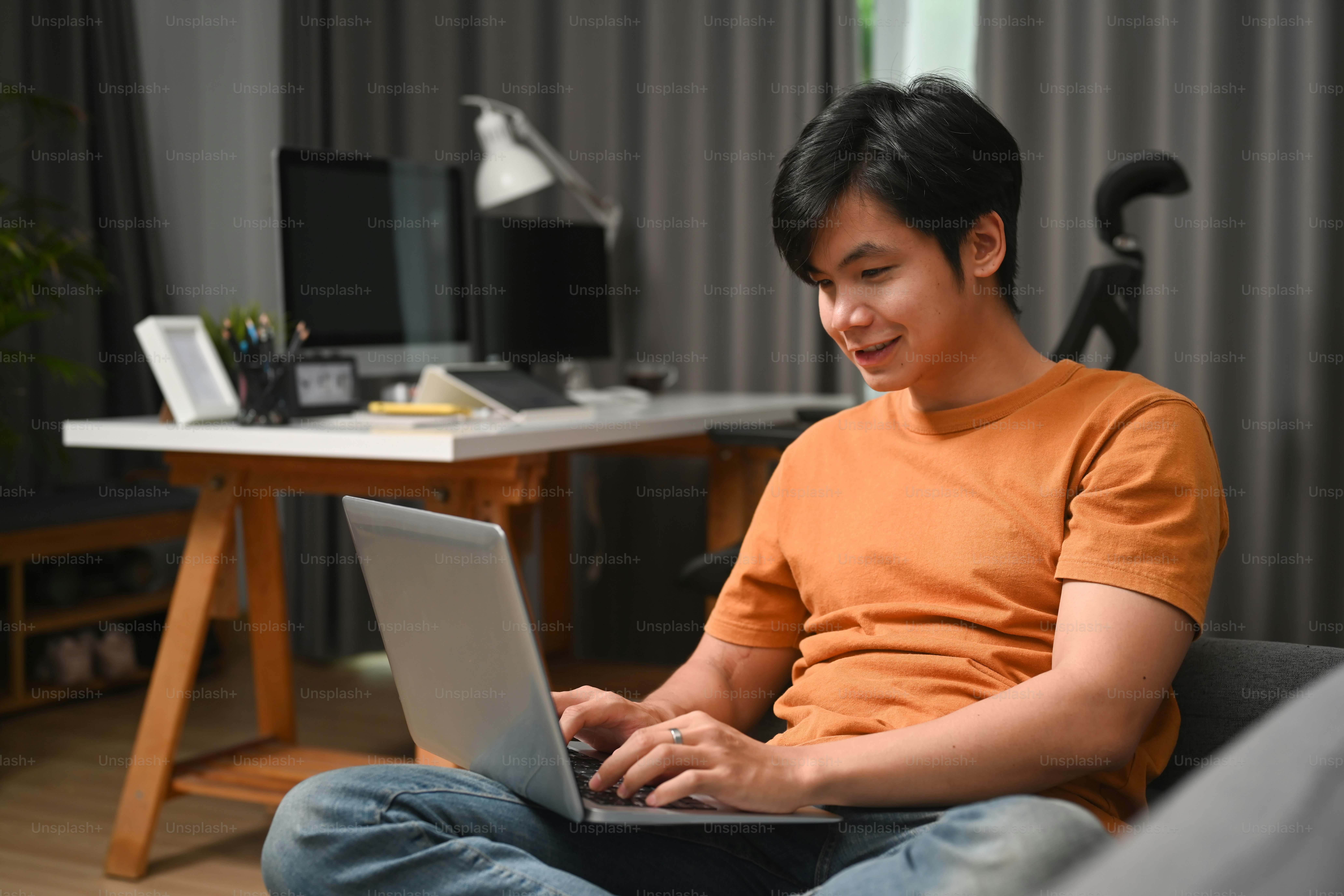 Pria kasual tersenyum duduk di sofa dan menggunakan komputer laptop.