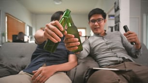 Two asian man drinking beer while sitting on sofa in living room.
