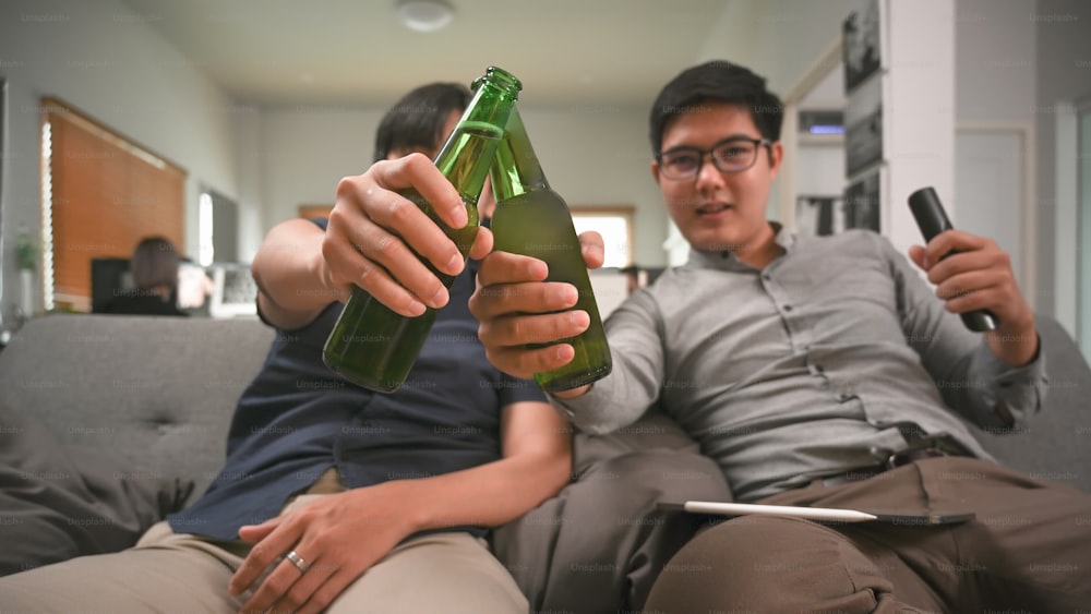 Due uomini asiatici che bevono birra mentre sono seduti sul divano in soggiorno.