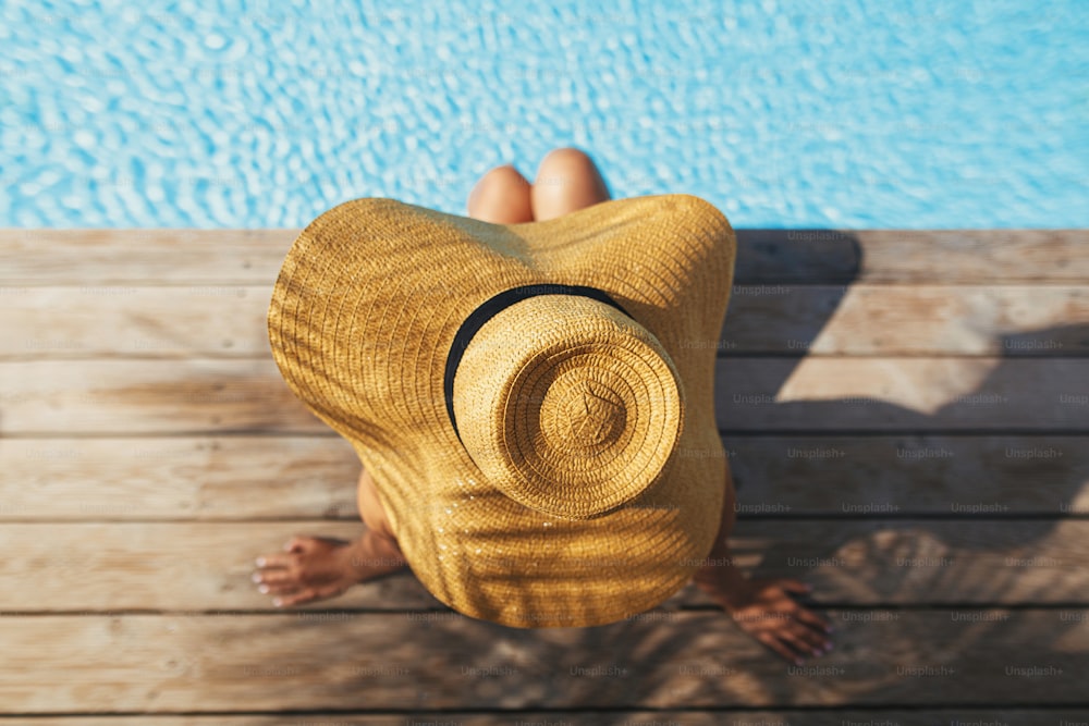 Beautiful woman in hat relaxing on wooden pier under palm leaves shadow at edge of pool, enjoying summer vacation. Top view of slim young female sunbathing at swimming pool. Holidays and travel