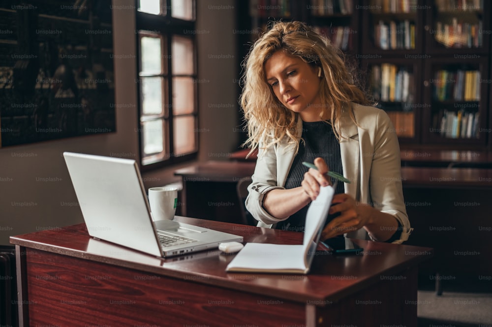 Jeune belle femme d’affaires blonde utilisant un ordinateur portable et un planificateur d’affaires tout en travaillant dans un bureau