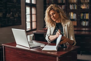 Junge schöne blonde Geschäftsfrau, die einen Laptop und einen Geschäftsplaner benutzt, während sie in einem Büro arbeitet
