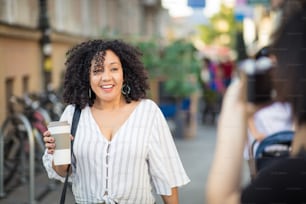 Mulher sorridente andando pela rua com xícara de café. Mulher tirando foto dela.