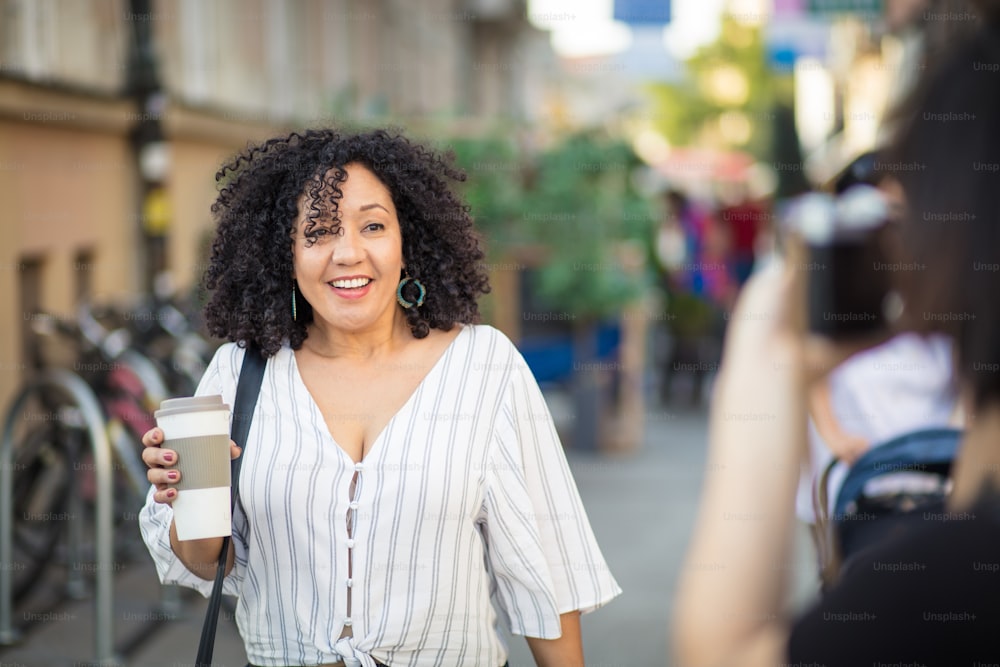 Smiling woman walking trough street with cup of coffee. Woman taking photo of her.