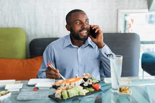 Lunch during working day. Business conversation. Young handsome afro american businessman eating sushi with sticks in a cafe, while talking phone and smiling