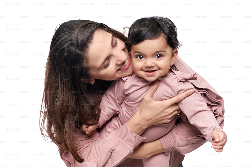 Portrait of cute mother with daughter having fun, isolated on white studio background. Young attractive woman hugging sweet adorable child, baby girl looking at camera, happy childhood concept.