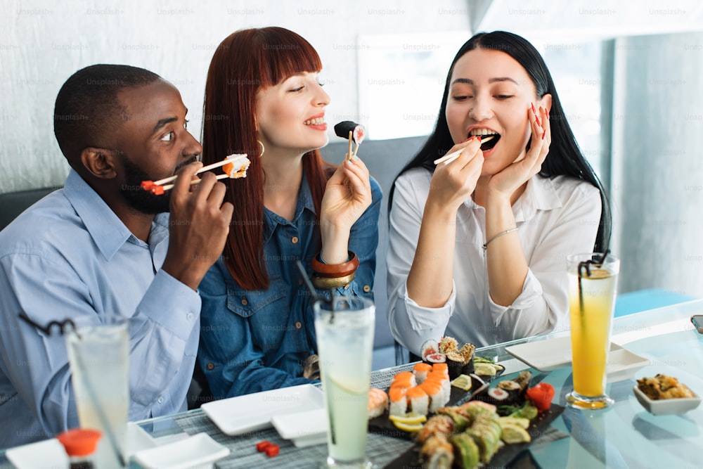 Gente, comida, diversión, comida de sushi. Primer plano de tres amigos multiétnicos emocionados, hambrientos y divertidos, divirtiéndose en el café comiendo comida asiática, tomando rollos de sushi con palitos de bambú cortados