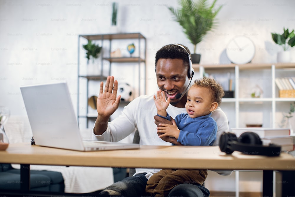 Afro american businessman with his little son on hands leading working meeting through video chat from home. Young father using modern laptop for work.