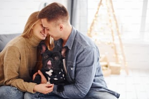 Happy young modern couple sitting on the sofa at home with christmas tree with their cute dog.