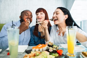 Giovani amici multirazziali allegri che mangiano sushi usando le bacchette, che sembrano positivi e felici, seduti in un ristorante asiatico e sorridono. Concetto di persone, emozioni, cibo e amicizia