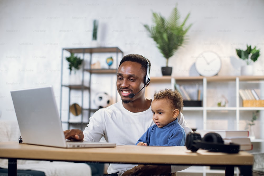 Afro american man in headset using modern laptop for work and taking care of his cute son. Male freelancer working from home with child on knees.