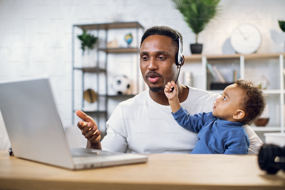 African man in headset having video chat on laptop while sitting at table. Young father trying to work remotely while taking care of cute baby boy.