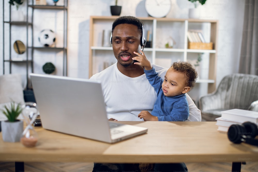 African man in headset having video chat on laptop while sitting at table. Young father trying to work remotely while taking care of cute baby boy.