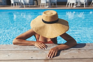 Beautiful woman in hat relaxing in water at pool wooden pier, enjoying summer vacation at tropical resort. Slim young female sunbathing at swimming pool edge, view above. Holidays and travel