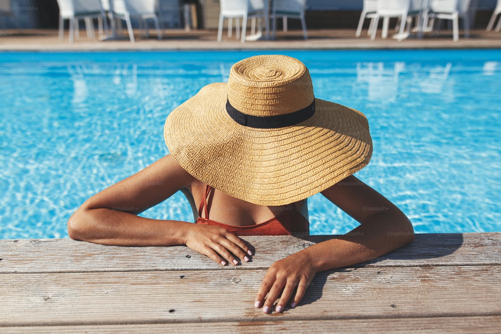 Beautiful woman in hat relaxing in water at pool wooden pier, enjoying summer vacation at tropical resort. Slim young female sunbathing at swimming pool edge, view above. Holidays and travel