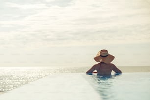 Beautiful woman in hat relaxing at edge in blue pool, enjoying summer vacation and calm sea view. Slim young female relaxing at tropical resort in swimming pool. Travel and Holidays. Space for text