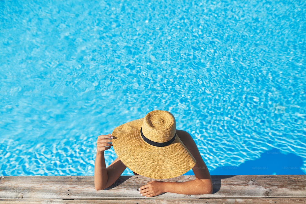 Enjoying summer vacation. Stylish woman in hat relaxing in pool water at wooden pier, top view. Slim young female sunbathing at  swimming pool edge. Travel and Holidays. Space for text