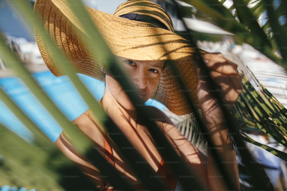 Enjoying summer vacation. Portrait of beautiful carefree woman in hat relaxing under palm leaves at tropical resort. Holidays and travel. Happy young female sunbathing at swimming pool.