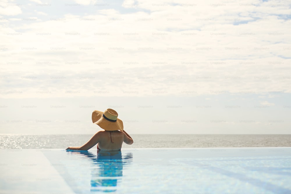 Hermosa mujer con sombrero relajándose en el borde de la piscina azul, disfrutando de las vacaciones de verano y la tranquila vista al mar. Mujer joven delgada que se relaja en el resort tropical en la piscina. Viajes y vacaciones. Espacio para el texto