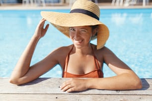 Beautiful happy woman in hat relaxing in water at pool wooden pier, enjoying summer vacation at tropical resort. Portrait of smiling young female sunbathing at swimming pool edge. Travel
