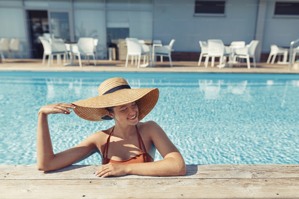 모자를 쓴 아름다운 평온한 여자는 수영장에서 휴식을 취하고 열대 리조트에서 여름 휴가를 즐기고 있습니다. 수영장 가장자리에서 일광욕을 하는 행복한 젊은 여성의 초상화. 여행 및 휴일