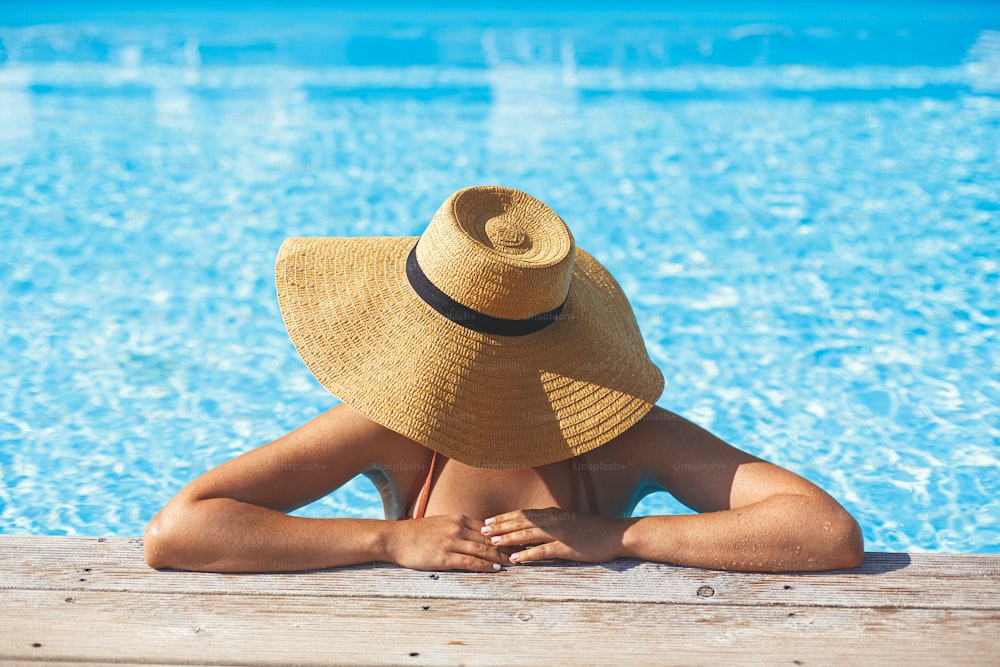 Beautiful woman in hat relaxing at pool wooden pier in water, enjoying summer vacation at tropical resort. Slim young female sunbathing at swimming pool edge, view above. Holidays and travel