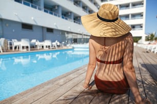 Beautiful woman in hat relaxing under palm leaves  shadow on wooden pier at pool, enjoying summer vacation. Slim young female tanning at swimming pool at tropical hotel resort