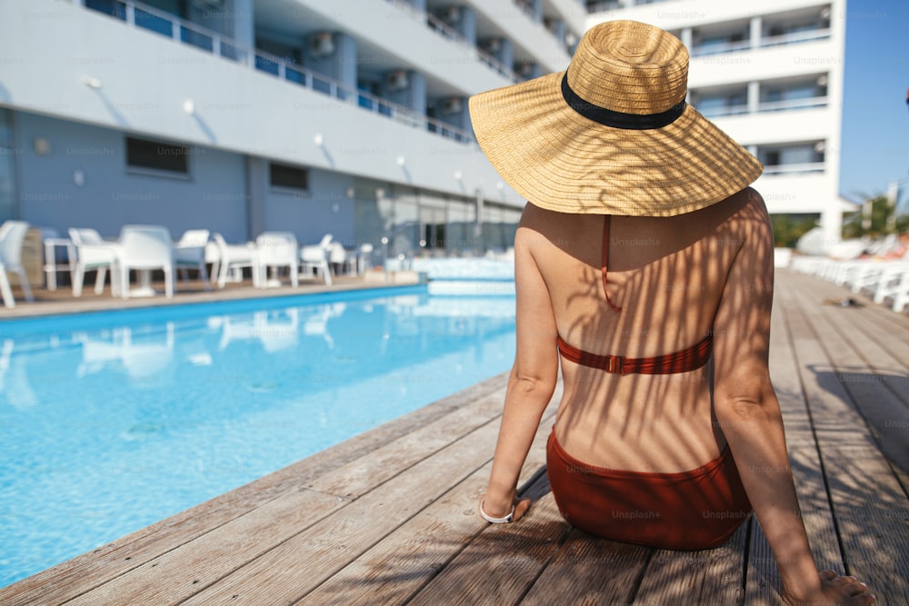 Schöne Frau mit Hut, die sich unter Palmen entspannt, hinterlässt Schatten auf dem hölzernen Pier am Pool und genießt den Sommerurlaub. Schlanke junge weibliche Bräunung im Schwimmbad des tropischen Hotelresorts