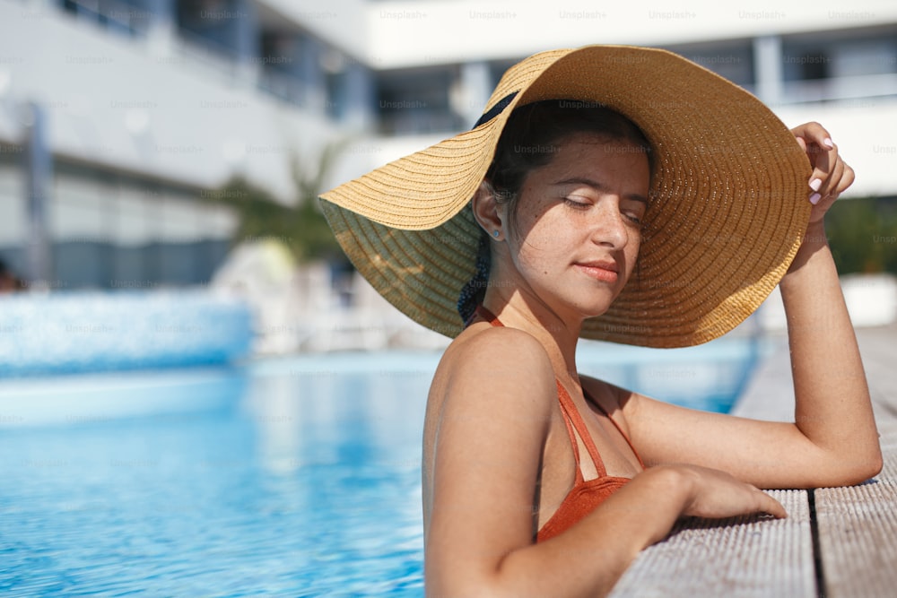 Beautiful calm woman in hat relaxing in pool water, enjoying summer vacation. Stylish slim young female sunbathing and resting at swimming pool edge at tropical resort. Holidays and travel
