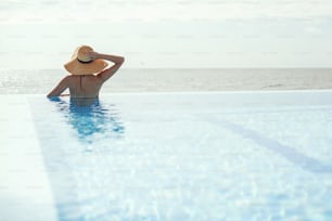 Vacanze estive. Bella donna in cappello che si rilassa sul bordo in piscina blu, godendo di una tranquilla vista sul mare. Giovane femmina magra che si rilassa al resort tropicale in piscina. Viaggi e vacanze. Spazio per il testo