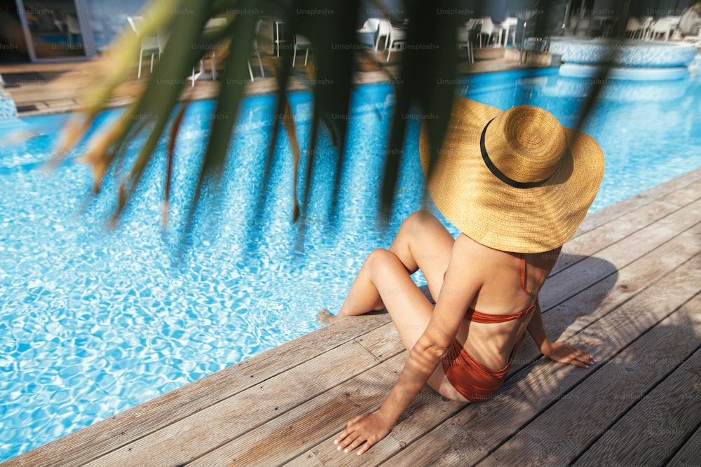 Beautiful woman in hat relaxing under palm leaves on wooden pier at pool enjoying summer vacation. Slim young female tanning at swimming pool at tropical resort. Travel and Holidays