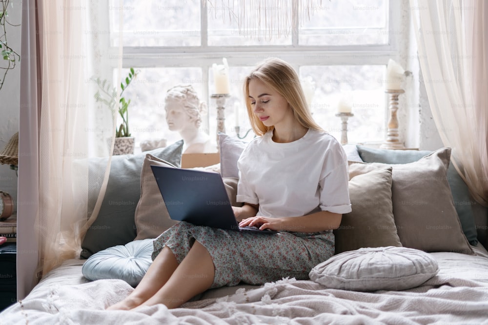 Joven mujer de cabello rubio sentada en la cama cómoda, usando una computadora portátil moderna, mirando la pantalla. Mujer que pasa la mañana en casa, trabajando en un dormitorio acogedor con un estilo interior bohemio